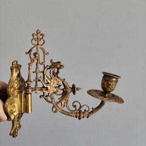 May include: Ornate gold-colored wall sconce with a candle holder. The sconce features intricate scrollwork and floral designs. The candle holder is a small cup shape with a decorative base. The sconce is mounted on a wall.