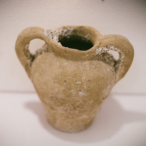 Puede incluir: Una pequeña maceta de terracota rústica con dos asas. La maceta tiene una superficie rugosa y texturizada y es de color marrón claro.