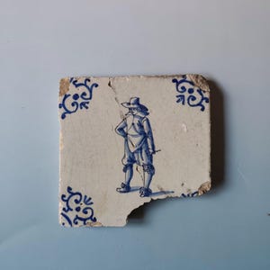 Puede incluir: Fragmento de baldosa de cerámica blanca con una ilustración azul de una persona con vestimenta histórica. La baldosa tiene diseños decorativos azules en las esquinas y un borde astillado, lo que sugiere edad y desgaste.