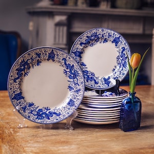 May include: A collection of white ceramic plates with a blue floral and dragon design. The plates are stacked and displayed on stands. A small blue glass vase holds a single yellow and orange tulip. The plates are on a wooden table.