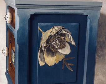May include: A blue painted wooden bedside table with a floral design on the front. The bedside table has two drawers and sits on four turned legs.