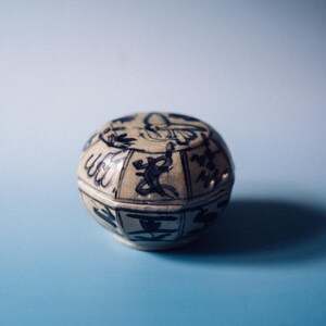 Peut inclure: Petite boîte à bijoux ronde en céramique avec couvercle. La boîte est blanc cassé avec des motifs bleus peints, dont des figures et des motifs stylisés. La boîte repose sur une surface bleue.