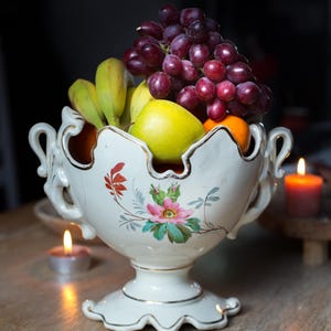 Könnte beinhalten: Eine weiße Porzellan-Obstschale mit floralem Muster und goldener Verzierung. Die Schale ist gefüllt mit Trauben, Bananen, Äpfeln und Orangen.