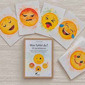 Könnte beinhalten: Ein Set aus 30 Gefühlskarten mit dem Titel Was fühlst du? mit handillustrierten Aquarell-Emojis. Die Schachtel zeigt fünf kleine Emojis und den Text 30 Gefühlskarten, Handillustrierte Aquarellmotive, Spielideen & Wissenswertes.
