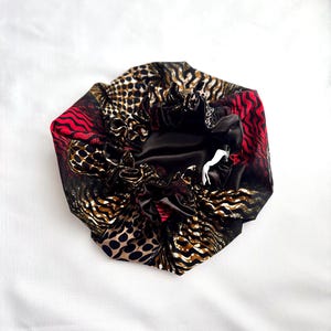 May include: A satin bonnet in black, red, and brown with an animal print. The bonnet has a black satin interior and an elasticated edge. The exterior features leopard and zebra patterns with red detailing.