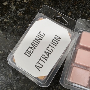 May include: Clear plastic clamshell packaging containing wax melts. The label reads "DEMONIC ATTRACTION" in bold, black text. The wax melts are a light pink color. The packaging is on a dark, textured surface.