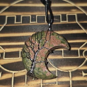 May include: A crescent-shaped pendant made of green and pink stone, wire-wrapped with a tree design. The pendant hangs from a black cord. The word "Astra" is visible in the lower left corner. The pendant is displayed on a wooden surface.