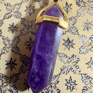 May include: A hexagonal, pointed amethyst crystal pendant featuring a deep purple hue with natural internal inclusions. The crystal is securely held by a polished gold-tone metal cap and bail, suspended from a dark cord necklace.