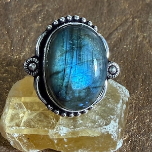 May include: A silver ring featuring an oval labradorite gemstone. The stone displays iridescent blue and green hues. The ring has a decorative silver setting with beaded and floral accents. The ring is displayed on a yellow-tan stone.