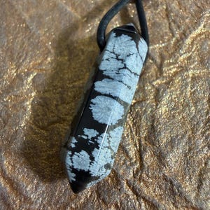 May include: A black and white snowflake obsidian crystal pendant. The polished stone is rectangular with a pointed end and is suspended from a black cord. The stone has a pattern of white snowflake-like inclusions.
