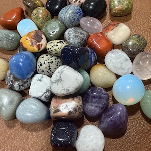 May include: A collection of polished gemstones in various colors and patterns. The stones include shades of blue, purple, red, green, and white. Some stones have speckled or marbled designs, while others are solid. The gemstones are smooth and rounded.