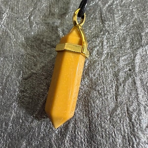 May include: A golden-yellow crystal pendant necklace. The crystal is a hexagonal shape with gold-coloured metal accents. The necklace has a black cord.