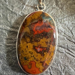 May include: An oval-shaped pendant featuring a polished, multi-colored stone with intricate patterns of deep red, earthy brown, and ochre yellow. The stone is encased in a smooth, silver-toned metal bezel setting with a matching silver-toned bail at the top.