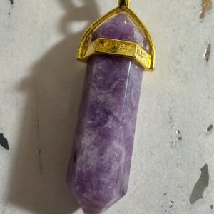 May include: A hexagonal, pointed purple crystal pendant suspended from a black cord. The crystal features a polished, marbled surface with varying shades of violet and white. It is secured by a gold-toned metal cap with a small loop at the top.