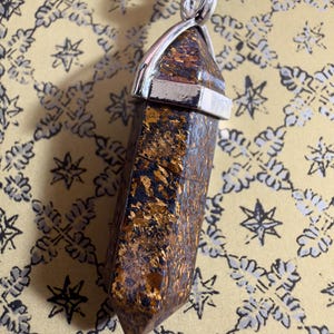 May include: A hexagonal point pendant made of brown and metallic gold stone, secured by a silver-colored metal cap. The pendant is suspended from a black cord, featuring a faceted, elongated shape with distinct dark and shimmering golden inclusions.
