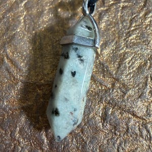 May include: A light green and black speckled crystal pendant with a silver-colored metal cap and a black leather cord. The crystal is a long, hexagonal shape. The background is a textured, gold-colored surface.