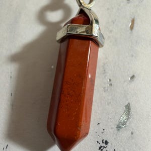 May include: A hexagonal point pendant made of smooth, opaque red jasper stone. The stone has a polished finish with subtle dark speckling. It is secured by a silver-toned metal cap with a bail at the top, attached to a black cord necklace.