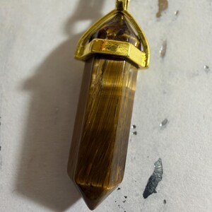 May include: A tiger's eye gemstone pendant shaped as a hexagonal prism with a pointed tip. The stone features brown and golden-yellow chatoyant bands. It is secured by a polished gold-tone metal cap and bail, suspended from a black cord necklace.