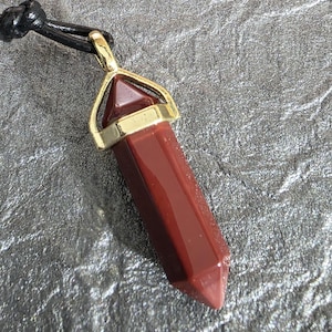 May include: A red gemstone pendant necklace with a gold-coloured metal setting. The pendant is a long, faceted shape, suspended from a black cord. The gemstone has a polished, glossy appearance.