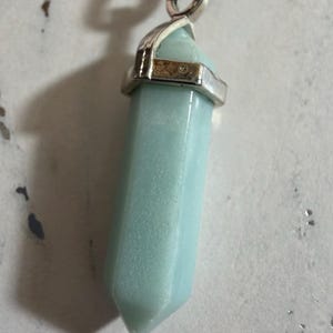 May include: A pale mint green, hexagonal, point-cut gemstone pendant suspended from a black cord. The stone is secured by a polished silver-toned metal cap with a circular bail at the top, creating a simple, vertical, geometric jewelry piece.