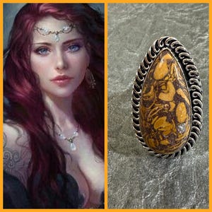 May include: A silver-toned ring with a teardrop-shaped, patterned stone in shades of brown and tan. The stone is surrounded by a twisted rope design. The ring is set against a textured gray background.