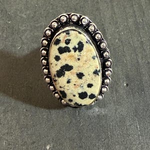 May include: Oval-shaped ring featuring a speckled stone with black spots and a silver-colored beaded border. The stone has a cream-colored base. The ring is set on a dark gray surface.