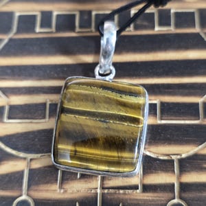 May include: A square tiger's eye pendant with a silver-colored bezel hangs from a black cord. The gemstone displays golden-brown bands. The word "Astra" is visible in the lower left corner. The pendant is displayed on a wooden surface.