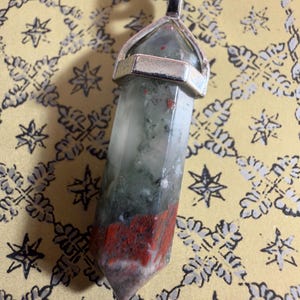 May include: A pointed, hexagonal crystal pendant featuring a blend of translucent white, mossy green, and opaque red mineral inclusions. The stone is secured in a silver-toned metal cap with a bail, suspended from a black cord necklace.