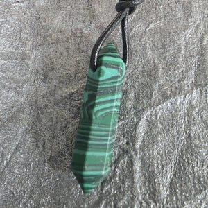 May include: A green and black malachite pendant with a leather cord. The gemstone is a rectangular shape with a pointed end. The stone has a striped pattern of dark green and black. The leather cord is black and knotted at the top.