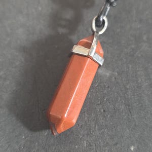 May include: A reddish-brown gemstone pendant necklace. The pendant is a hexagonal shape with silver-colored metal accents. The necklace cord is black and knotted. The word "Astra" is in the bottom left corner.
