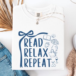 May include: White sweatshirt with the words "READ RELAX REPEAT" in blue script. The design includes illustrations of a book, seashell, strawberry, and other leisure items. A silver necklace and blue jeans are also visible.