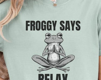 May include: A light green t-shirt with a black graphic of a frog in a yoga pose. The text "Froggy Says Relax" is printed in black on the shirt.