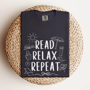May include: A navy blue t-shirt featuring the phrase "READ RELAX REPEAT" in white, accompanied by illustrations of a sun, book, beach items, and a cocktail. The shirt is folded on a woven mat.