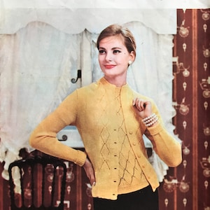 May include: A yellow knitted cardigan with a diamond pattern and a mock neck. The cardigan is buttoned up and has long sleeves. The model is wearing a black skirt and a pearl bracelet.