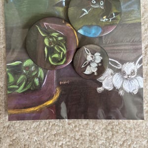 Könnte beinhalten: Set mit drei runden Ansteckbuttons mit Fantasy-Kunst-Designs. Ein Button zeigt ein grünes Wesen, ein anderer ein weißes und silbernes Wesen und der dritte ein schwarzes Wesen. Die Buttons befinden sich auf einer bedruckten Karte mit passendem Artwork.