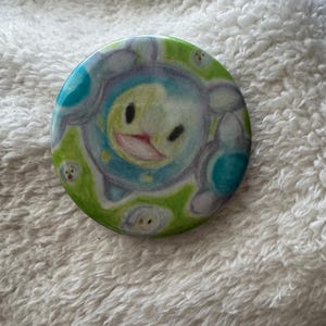 May include: A round button pin with a cartoon character design. The pin features a central character with a smiling face, surrounded by a blue and green pattern. Small, similar characters are scattered around the edge.