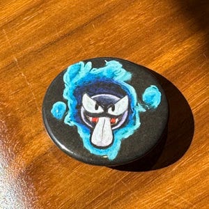 Könnte beinhalten: Ein runder, schwarzer Knopf mit einem Cartoon-Geist-Design. Der Geist hat eine weiße Zunge, einen roten Mund und weiße Augen, umgeben von blauen Flammen. Der Knopf liegt auf einer Holzoberfläche.