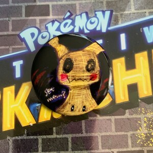 Könnte beinhalten: Ein runder Knopf mit schwarzem Hintergrund und einem Cartoon-Bild eines braunen und gelben Pokemon-Charakters mit roten Wangen. Der Charakter hat schwarze Augen und einen schwarzen Mund. Der Text "See me...?" ist in Weiß auf dem Knopf geschrieben.