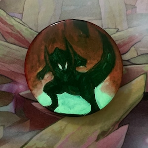 May include: A black and green glow-in-the-dark pin with a silhouette of a creature with horns and glowing eyes. The creature is set against a red and orange background.