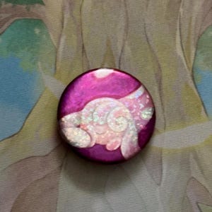 Puede incluir: Un botón redondo de color magenta con un diseño de ala blanco e iridiscente. El botón tiene un acabado brillante y probablemente está hecho de metal o plástico. El diseño del ala es detallado y parece estar pintado a mano.