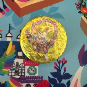 May include: A round, yellow button with a holographic effect featuring a cartoon rabbit character holding a weapon. The button has a purple and pink border and is set against a colorful, patterned background.