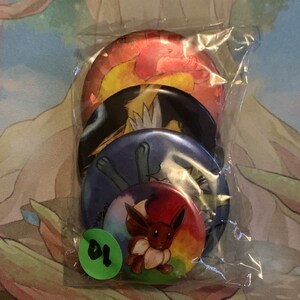 May include: Four round, colorful buttons featuring Pokemon characters. The buttons are in a clear plastic bag. The top button features a red and yellow Pokemon character. The second button features a black and white Pokemon character. The third button features a blue and white Pokemon character. The bottom button features a brown Pokemon character with a rainbow background.