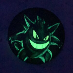May include: A black and green glow-in-the-dark illustration of a cartoon ghost with a mischievous grin. The ghost has a spiky, jagged outline and is set against a dark blue background.