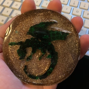 May include: A round, clear resin coaster with a gold glitter background and a green silhouette of a dragon.