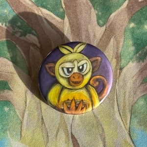 Könnte beinhalten: Ein runder Button mit einem Cartoon-Affen mit gelbem Fell und braunem Gesicht vor einem violetten Hintergrund. Der Affe hat einen ernsten Gesichtsausdruck. Es ist wahrscheinlich ein Sammlerstück oder ein Accessoire.
