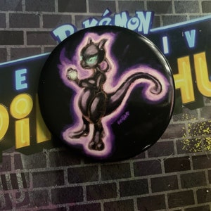 Könnte beinhalten: Ein schwarzer Knopf mit einer lila und weißen Illustration eines Pokémon-Charakters, "Mewtwo", auf schwarzem Hintergrund. Der Knopf hat einen weißen Rand und der Text "Pokémon Live" ist im Hintergrund sichtbar.