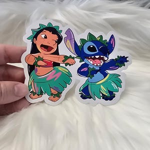 Puede incluir: Dos pegatinas coloridas con Lilo y Stitch de la pel&iacute;cula de Disney Lilo & Stitch. Lilo lleva una falda de hierba verde con detalles rosas y azules. Stitch lleva una falda de hierba azul con detalles verdes y amarillos. Ambos personajes est&aacute;n bailando.