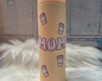 May include: A pale yellow reusable tumbler with a pink "MOM" graphic and multiple white coffee cup stickers. The tumbler has a straw.