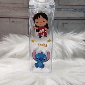 Puede incluir: Botella de agua rectangular transparente con los personajes de dibujos animados Lilo y Stitch. La botella tiene una tapa transparente y la palabra "ohana" impresa en marr&oacute;n. Los personajes son a color y la botella est&aacute; sobre una superficie blanca y esponjosa.