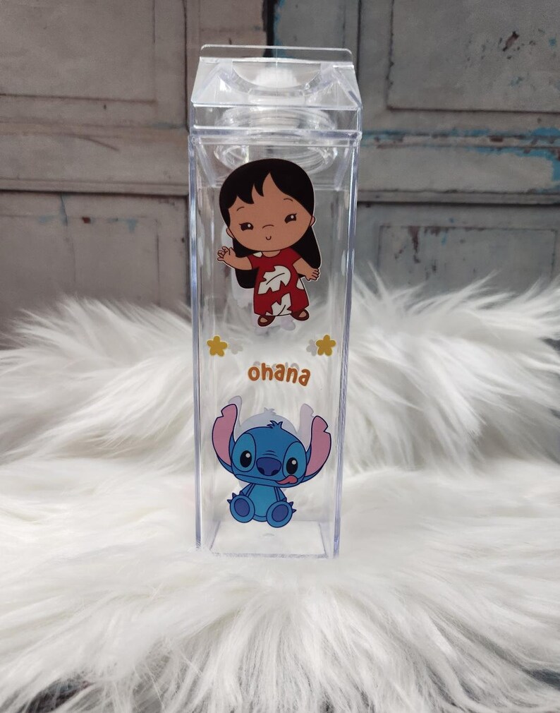 Puede incluir: Botella de agua rectangular transparente con los personajes de dibujos animados Lilo y Stitch. La botella tiene una tapa transparente y la palabra "ohana" impresa en marr&oacute;n. Los personajes son a color y la botella est&aacute; sobre una superficie blanca y esponjosa.